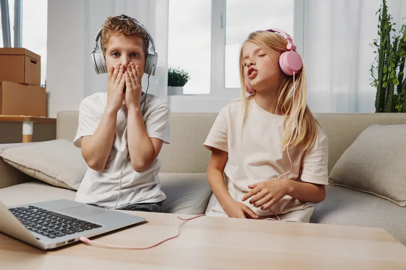 Two children with headphones enjoy music at home, sitting on a sofa with a laptop nearby. The boy looks surprised while the girl sings along, creating a playful atmosphere.