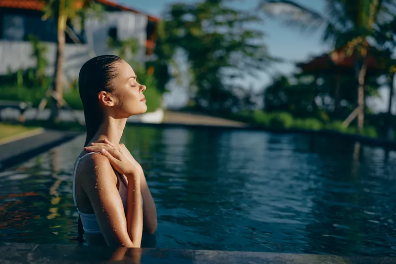 A young woman in a swimming pool enjoys a peaceful moment with eyes closed and hands on shoulders. The outdoor setting includes tropical plants and a sunny sky. Lifestyle concept.