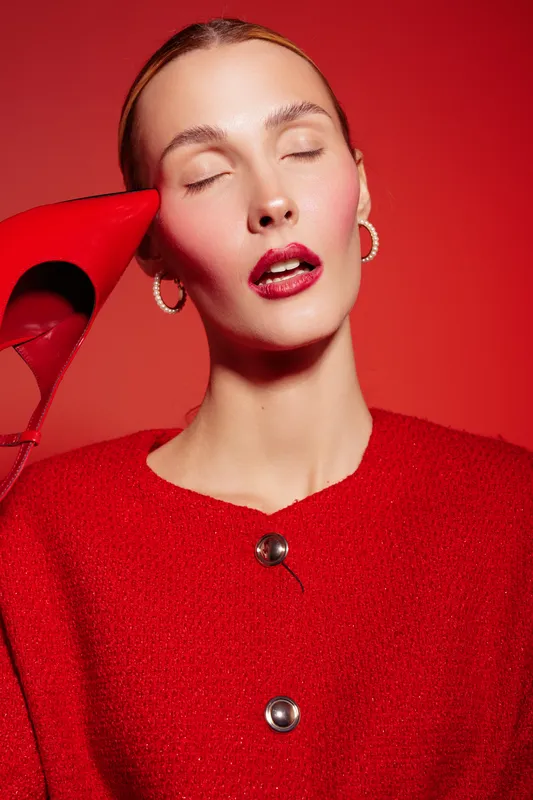 Close-up portrait of a woman with closed eyes wearing red lipstick and red sweater holding a red high heel shoe near her face. Concept of fashion, beauty, and style in studio.