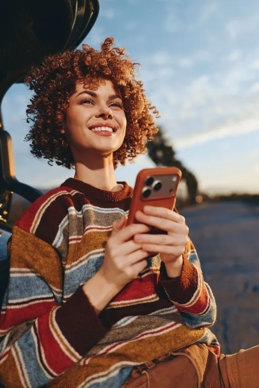 Smiling woman with curly hair holding smartphone outdoors, wearing striped sweater, standing by road at sunset, lifestyle travel, casual fashion portrait, warm golden light