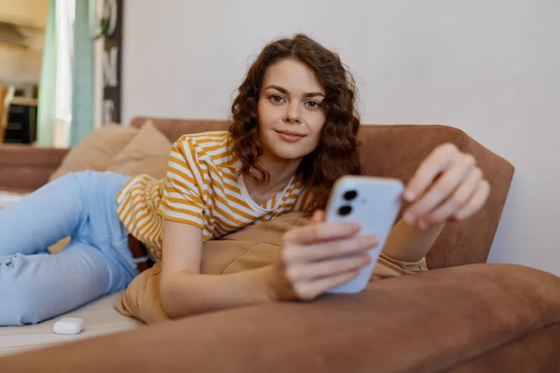 A woman lounges on a couch at home, taking a selfie with a smartphone. Casual lifestyle scene with striped shirt, cozy decor, and warm daylight creating a relaxed, authentic mood.