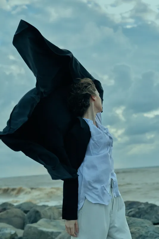 Wind catches flowing cloak on a woman at a rocky beach, dramatic movement against cloudy sky and sea horizon, fashion powered by wind and ocean breeze