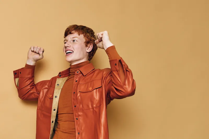Young man, smiling and celebrating with raised fists indoors against brown background. Casual dressed male with red hair in jacket, expressing happiness and excitement in modern studio portrait.