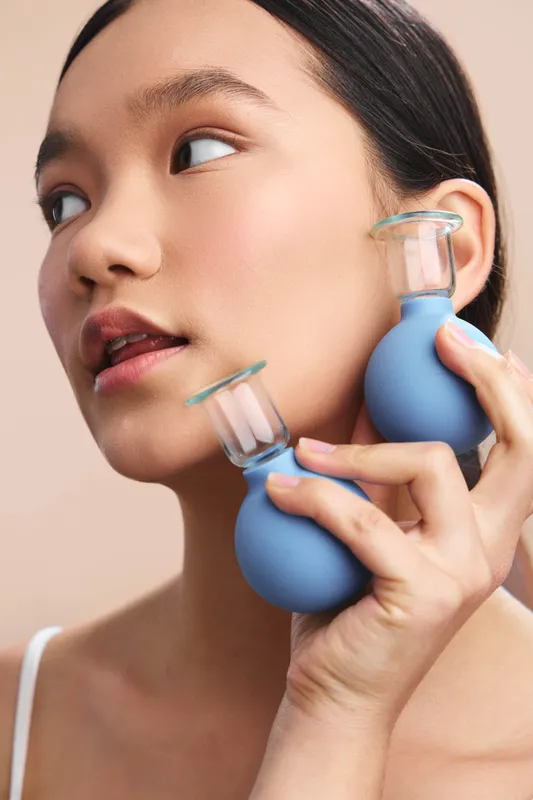 Asian woman with calm expression holds blue suction bulbs near her face in a soft studio shot, creating a focused, modern wellness vibe with simple lighting and gentle color tones.