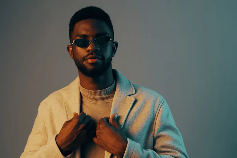 Stylish young African man in sunglasses and a sweater posing confidently against a pastel background, showcasing modern fashion and self expression