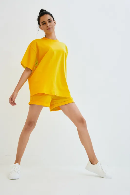 Young woman wearing bright yellow oversized t-shirt and shorts standing with wide legs on white background, casual summer outfit and confident pose in studio lighting.