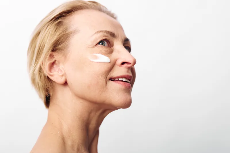 Mature woman with short blonde hair applies white facial cream on cheek, looking up and smiling. Concept of skincare, aging skin care, beauty routine, and self-care.
