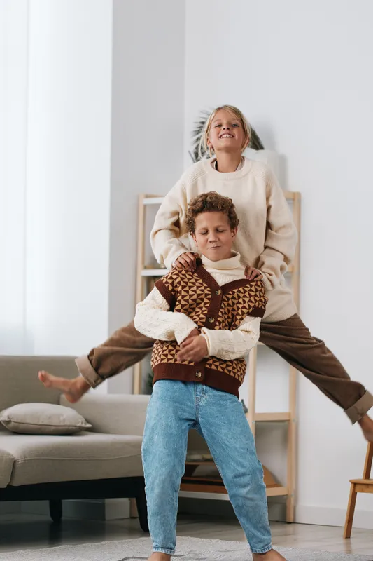 Two children play indoors in a joyful moment, one standing behind the other with arms around his shoulders; cozy knitwear, bright casual setting, and carefree energy.
