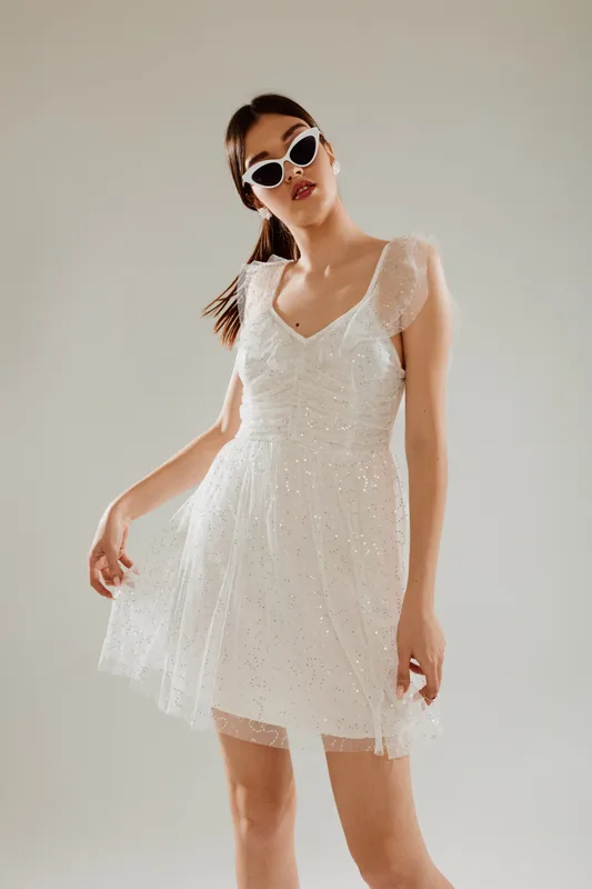 Young woman in a white lace dress and sunglasses poses confidently against a neutral background. Fashion concept highlighting beauty, style, and modern elegance.