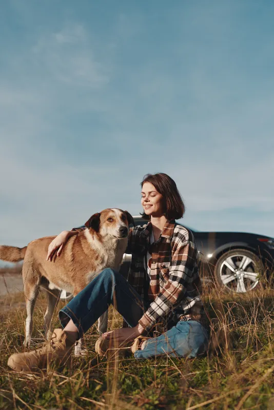 Woman and dog outdoors in a sunny field, sitting on grass with a friendly canine for companionship, wearing a plaid shirt, relaxed pose near a car, capturing simple countryside leisure.