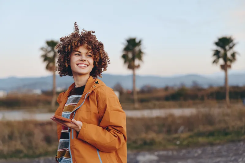 Woman lifestyle outdoors, smiling with curly hair, wearing orange jacket and denim, standing near palm trees on sunny landscape, casual travel mood, natural light.