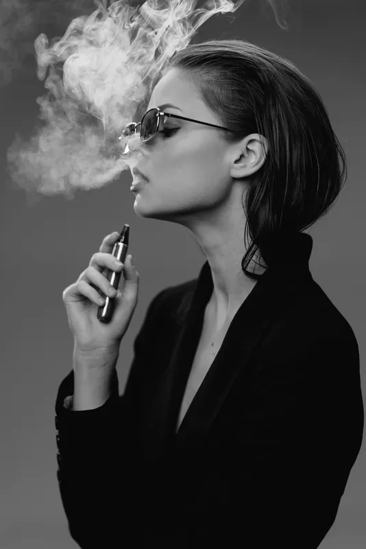 Black and white profile of a woman vaping with sunglasses exhaling smoke in dark outfit and short hair