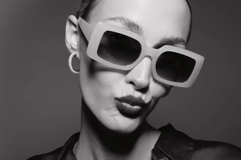 Close-up portrait of white woman with pale skin wearing large sunglasses and hoop earrings in black and white studio photo.