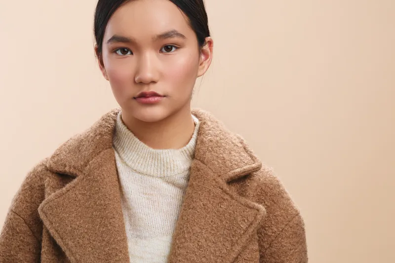 Asian woman wears a warm beige coat in a clean studio setting, presenting a calm, confident mood. Her composed expression and soft lighting convey elegance and approachable professionalism.