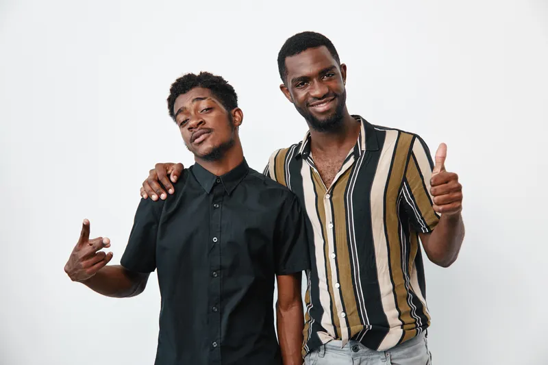 Two african men with dark skin posing together smiling and showing thumbs up and hand gesture on white background. Friends, casual clothing and positive mood.
