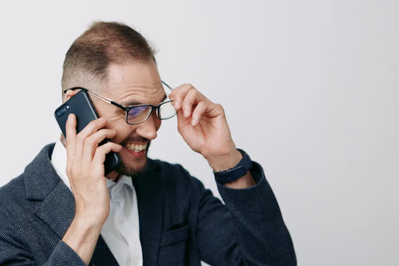 Professional businessman on a phone call, wearing a blazer and glasses, with a confident smile, isolated on a light colored background, conveying focus and energy.