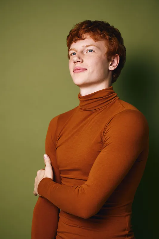 Portrait of confident young man with red hair wearing brown turtleneck sweater standing against green background looking sideways with calm expression and crossed arms.