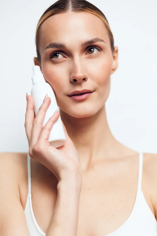 Smiling woman applying lotion from a bottle to her face. Close-up on a white background. Concept of skincare, beauty, and self care. Soft studio lighting creates a calm, fresh mood.