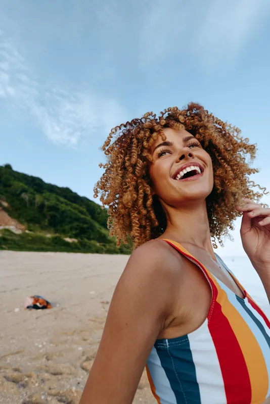 Happy woman with curly hair wearing colorful striped swimsuit smiling outdoors on a sunny beach. Blue sky and green hills in background create a joyful summer mood.