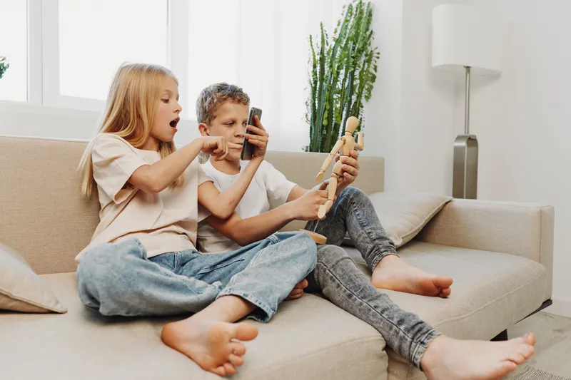 Two children sitting on a beige couch playing with wooden toys and a smartphone in a bright living room with natural light and houseplants in the background.