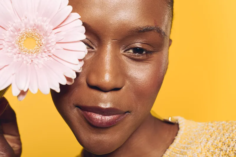 Close-up of a smiling African woman with dark skin holding a pink flower near her face against a yellow background. Concept of beauty, skincare, nature, and wellness.