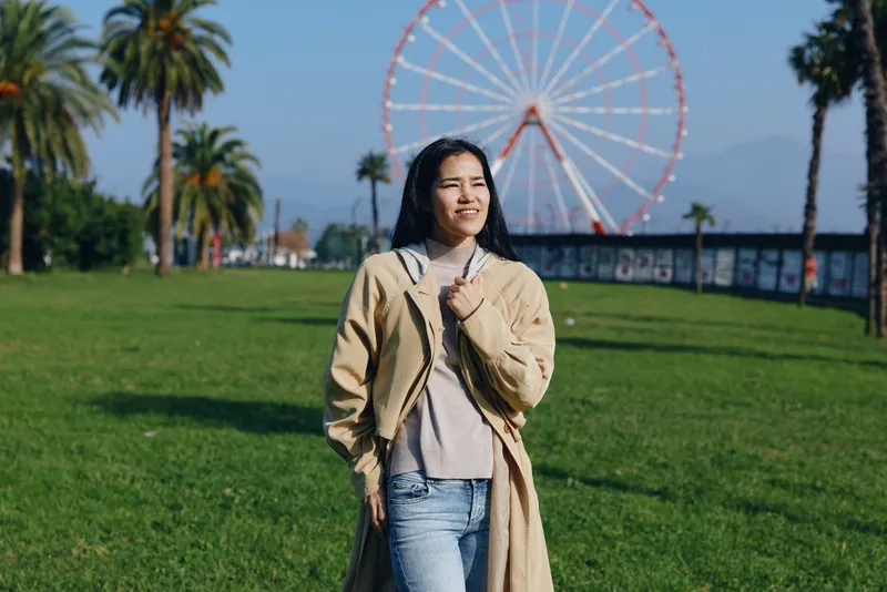 Woman in a beige coat and sweater stands in a sunny park, with a large ferris wheel in the background, conveying casual fashion and outdoor leisure on a bright day