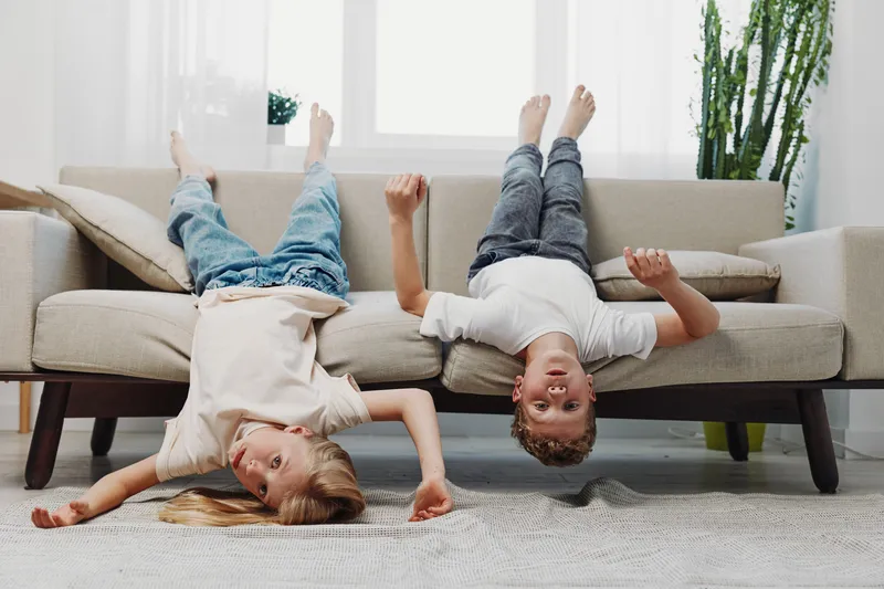 Two playful children lying upside down on a beige sofa in a bright living room. The joyful kids wearing casual clothes enjoy fun and carefree moments at home with natural light.