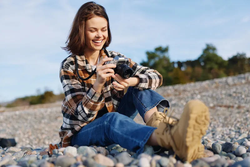 Photographer woman outdoor beach camera casual lens, smiling while adjusting the camera on a pebbled shore, enjoying sunny nature and a relaxed creative moment.