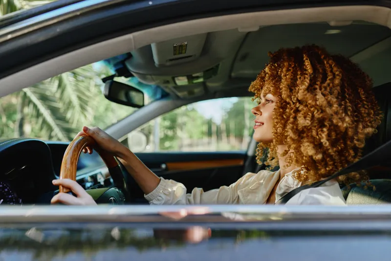 woman driving car with curly hair during daytime, natural light, casual clothing, relaxed and focused expression, green blurred background outside the window.