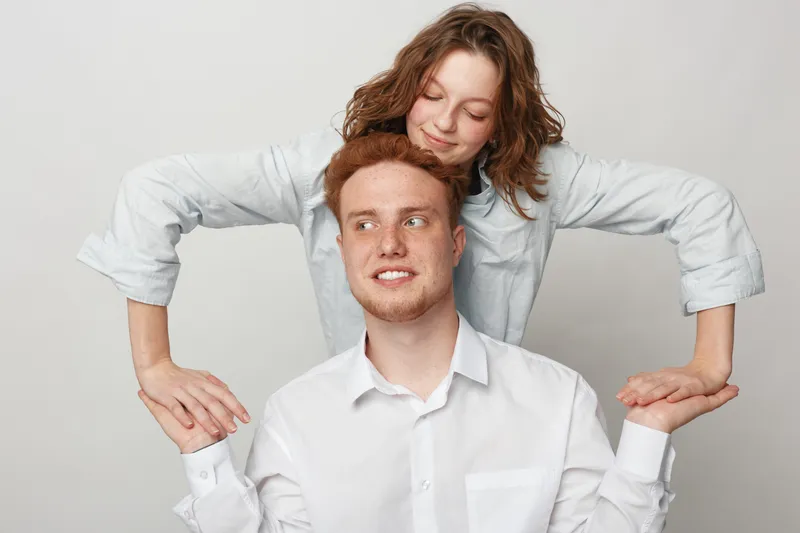 Young man in white shirt smiles while woman with curly hair stands behind him holding his hands. Studio portrait on light background with casual clothing and friendly mood.