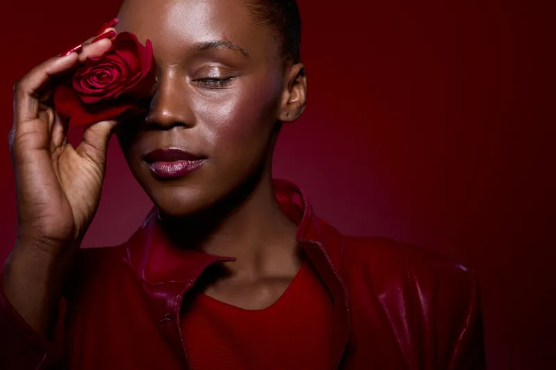 Close-up of a dark skinned African woman with eyes closed holding a red rose near her face. She wears red clothing against a dark red background. Concept of beauty, elegance, and romance.