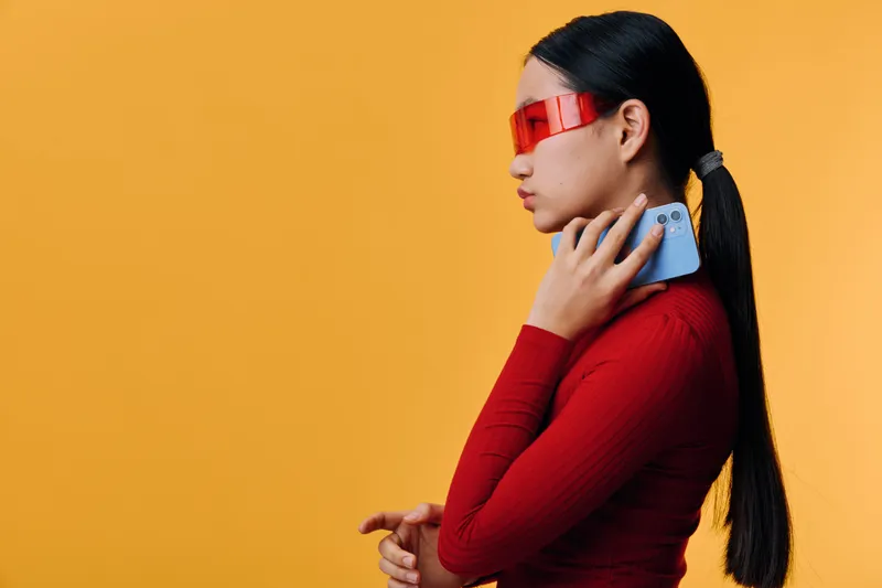 Woman with black hair wearing red turtleneck and futuristic red glasses holding smartphone near face on yellow background in studio. Profile view with casual modern style and tech gadget.
