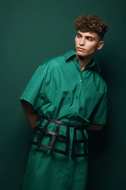 Young man with curly hair wears green oversized shirt and belt accessory posing on dark green background fashion stylish model trending beauty portrait confident young person.