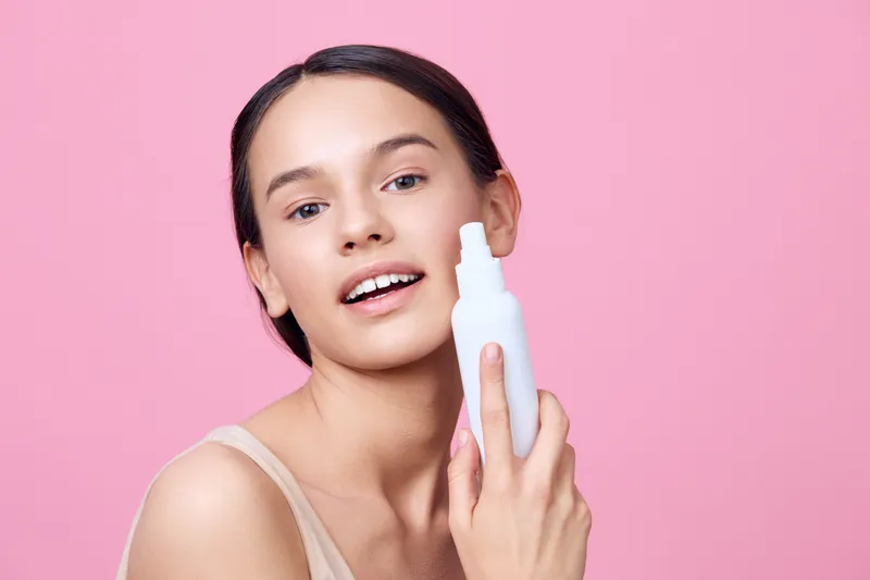 Young woman demonstrating skincare routine with a white spray bottle against a pink background, highlighting beauty and health