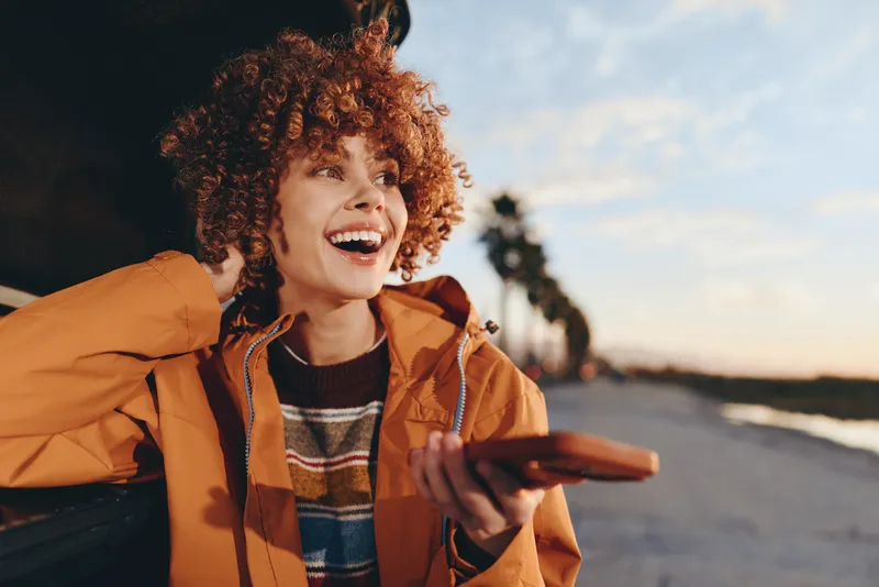 woman lifestyle sunset curly smile jacket road walking orange portrait, laughing curly haired woman in orange jacket and striped sweater walks on quiet road at sunset, warm light