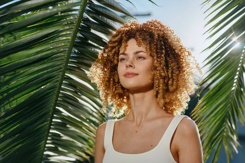 young woman with curly hair stands confidently under bright sunlight surrounded by tropical palm leaves, wearing a white tank top with natural makeup.