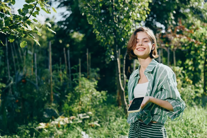 A smiling woman stands in a sunlit garden, holding a tablet, wearing a striped shirt and casual jacket.