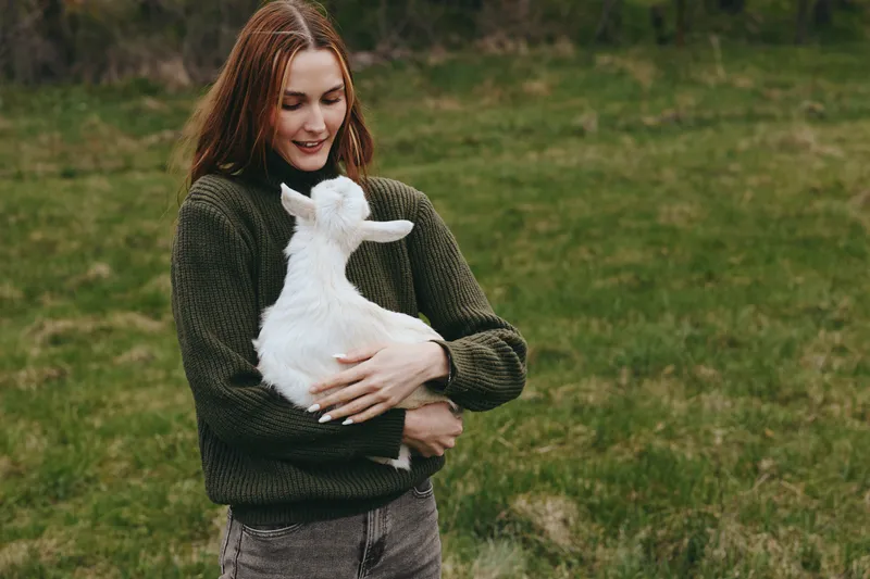 Young woman with long red hair wearing a green sweater gently holds a white baby goat in her arms outdoors on a grassy field, expressing warmth and care.