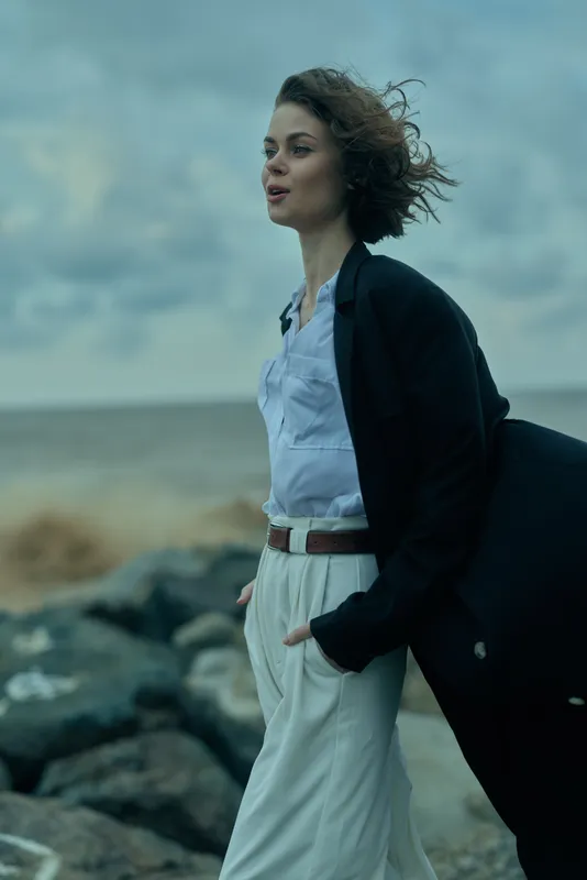 Woman walks along the windy coast in a light suit, blazer over shoulder, sea breeze, coastal landscape, fashion moment, sea spray and dramatic sky, confident stride