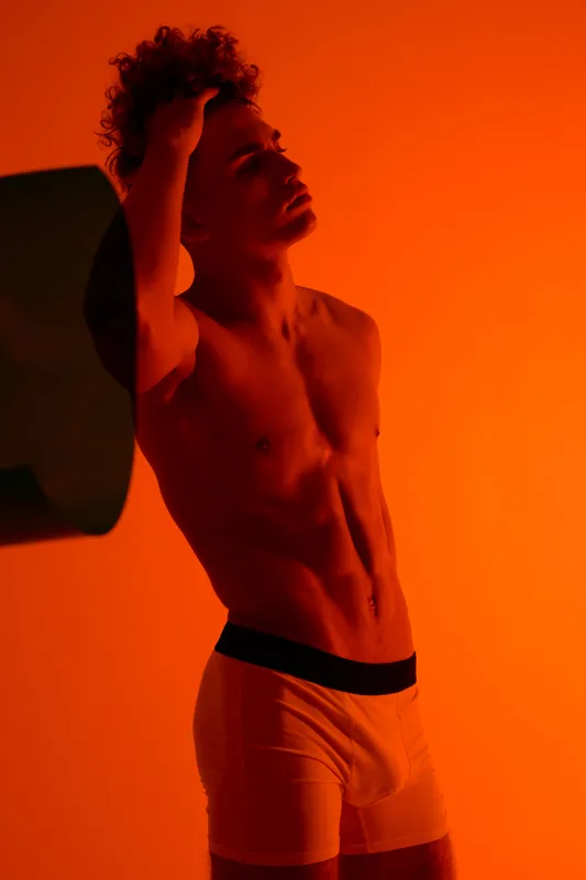 Young man with toned body standing faces sideways with hand on head wearing white briefs in orange studio lighting for fitness, health and muscle posing concept.