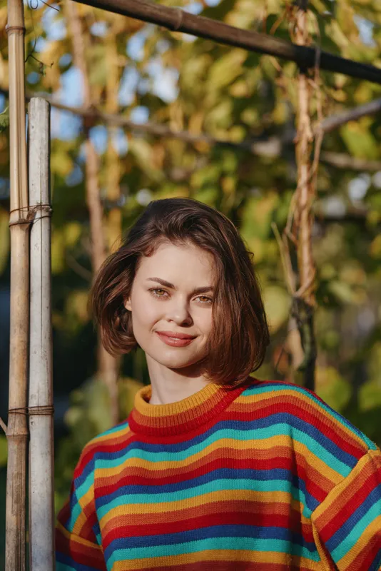 Woman, portrait, striped sweater, smiling, short hair, outdoor, garden, youth — Early twenties female in a colorful rainbow knit leaning on a bamboo pole, relaxed smile in warm natural light