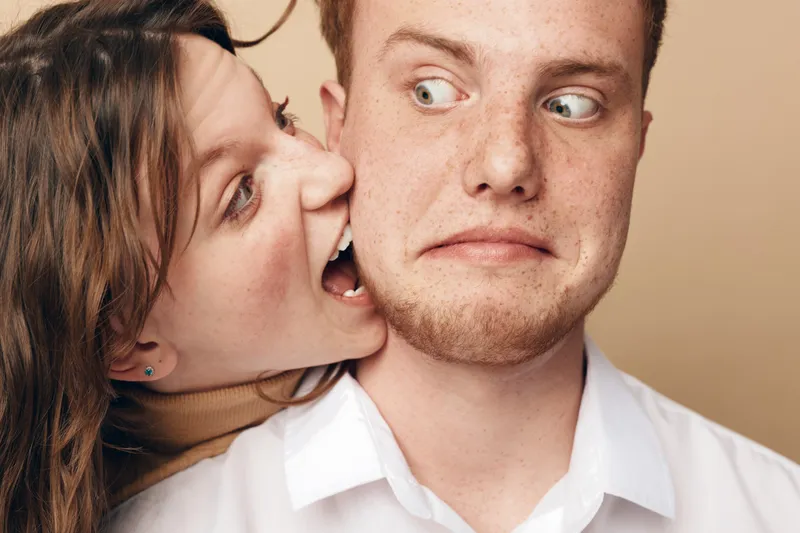 Young woman playfully bites young man on cheek while he looks surprised. Close-up portrait on beige background. Concept of fun, humor, relationship, and playful interaction between couple.