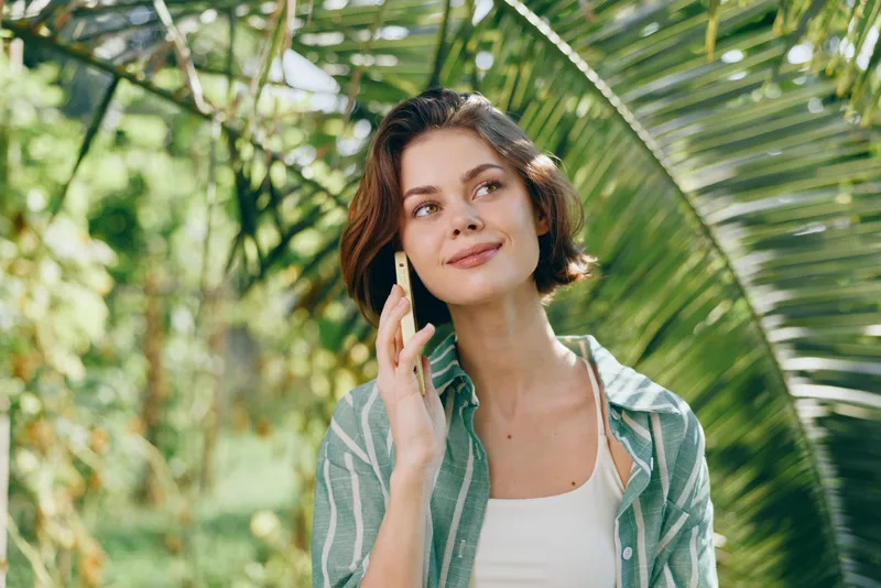 woman, outdoor portrait, cheerful, smiling, talking on phone, casual outfit, natural light, lush greenery background, garden setting, relaxed mood, confident expression, modern lifestyle vibe