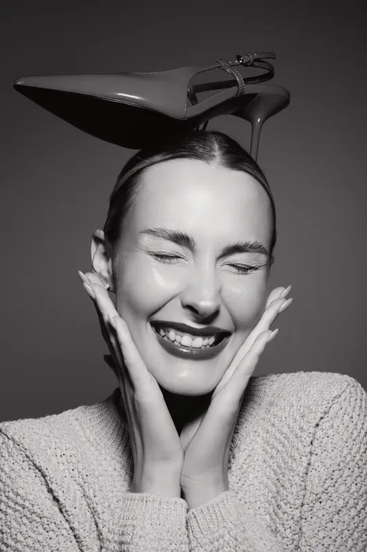 Happy white woman with light skin and closed eyes smiles joyfully with high heel shoe on her head in black and white studio portrait.