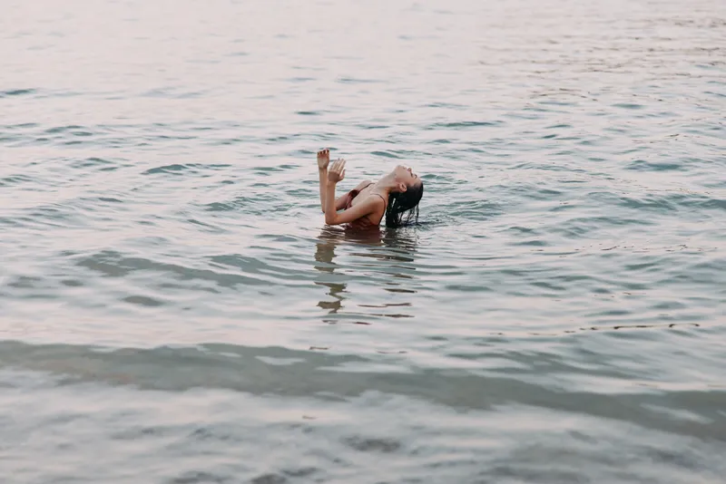 A solitary figure floats peacefully in calm water, partially submerged with arms raised, creating a serene focal point among gentle ripples and soft light.