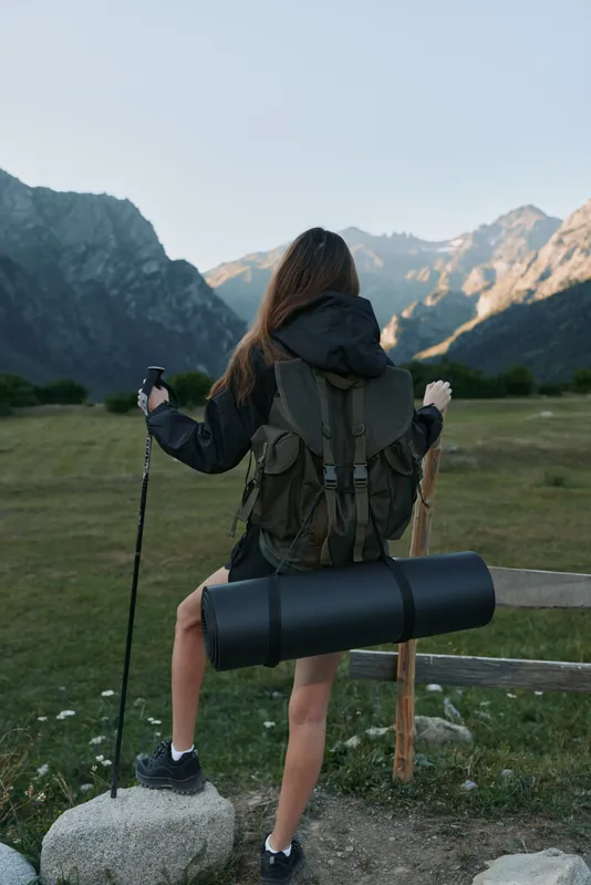A lone hiker stands with a large backpack in a rugged mountain landscape. The figure faces away, ready for a trek as warm light bathes the valley and peaks.