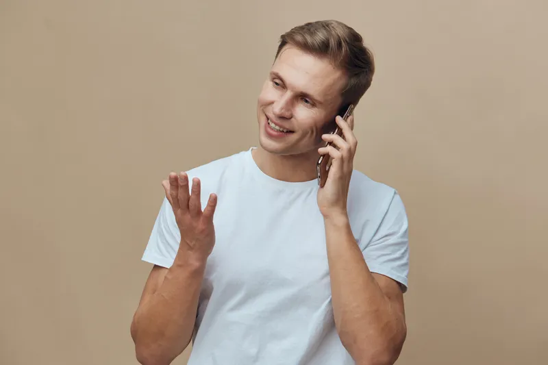 Smiling man talking on mobile phone holds hand raised and gestures casually light background. Young happy male using mobile device for communication or conversation in studio setting. People lifestyle
