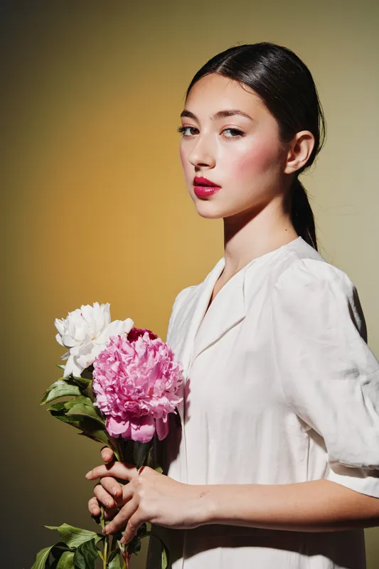 Young woman with minimal makeup and slicked hair holding pink and white peonies against a warm beige background. Artistic beauty-fashion photo emphasizing natural elegance.