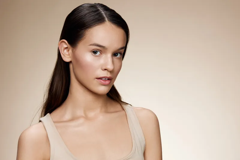 Portrait of a young woman with long hair wearing a light tank top against a soft beige background, conveying beauty and confidence
