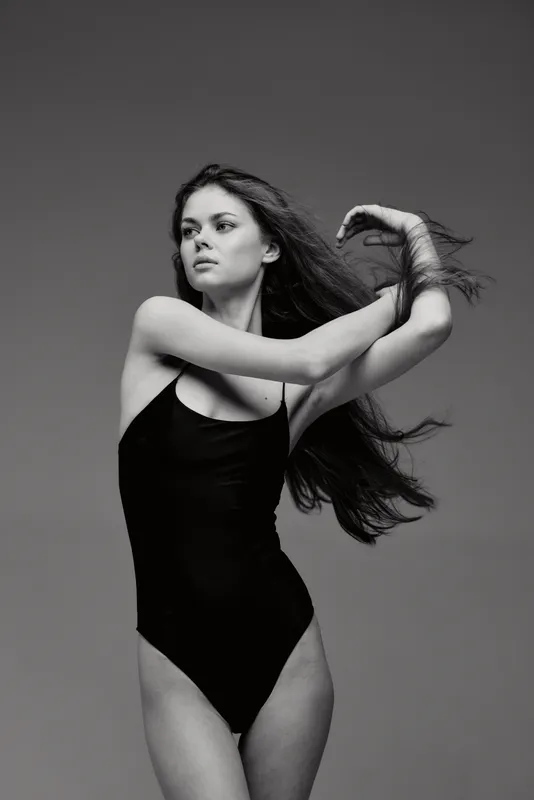 Elegant black and white woman posing in black swimsuit with flowing hair, expressing grace and confidence against simple gray background.